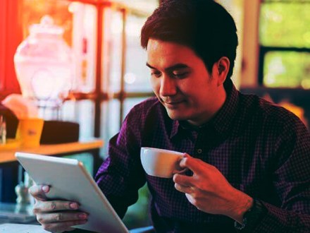 Smiling man reading his tablet while drinking coffee.