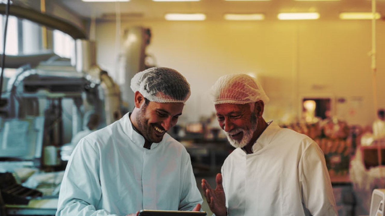 Two cheerful male colleagues in sterile clothes standing in food factory and looking at sales documents on digital tablet.