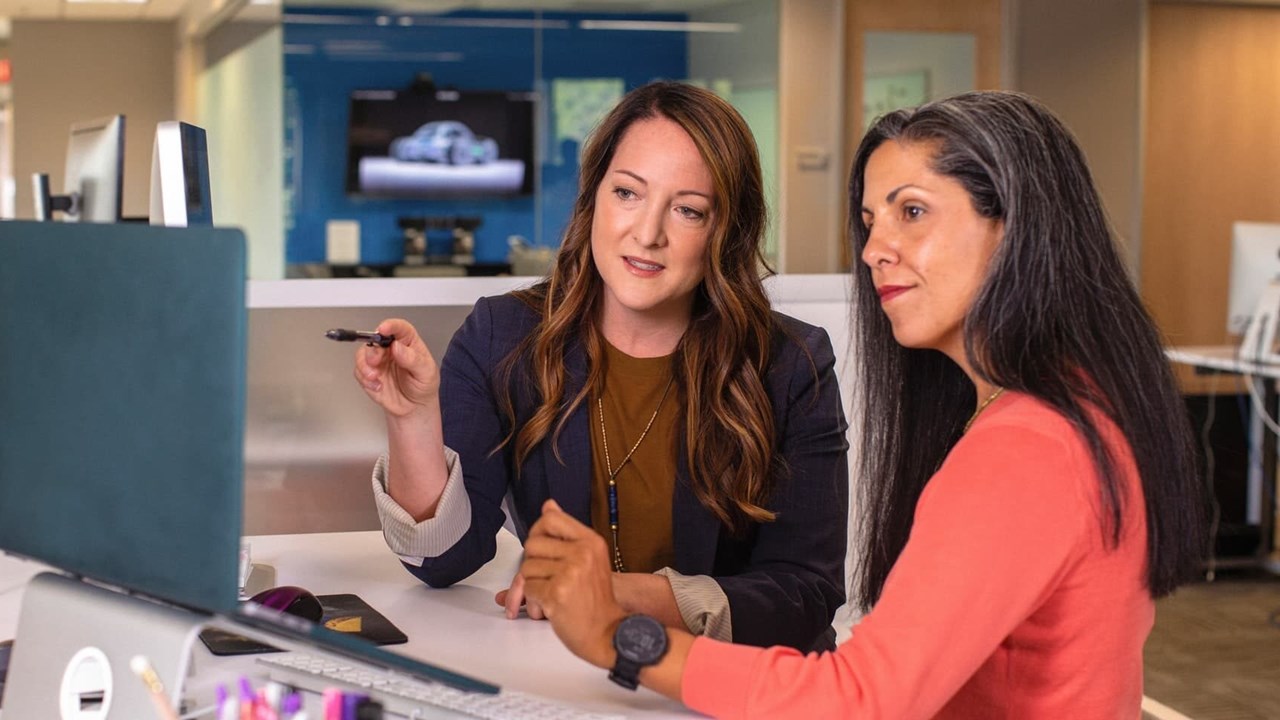 Two Business Women Talking About Sales In Office At Desk With Laptop