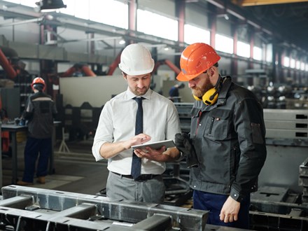 Young master in hardhat and bearded engineer discussing technical sketch on display of tablet in factory workshop.