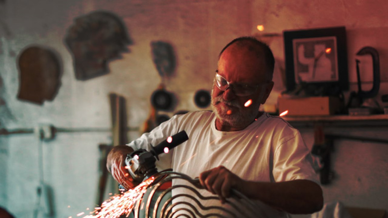 a Senior man using an angle grinder to create sculptures out of metal in his art studio.