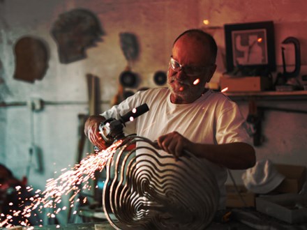 a Senior man using an angle grinder to create sculptures out of metal in his art studio.