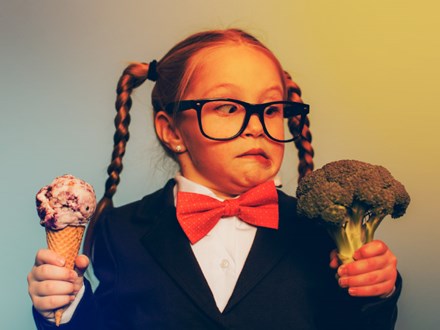 A young female nerd dressed in bow tie and eyeglasses is deciding between eating an ice cream cone or broccoli. She is making a disgusted face at the broccoli. She is choosing the treat.